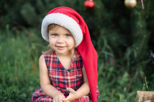 little girl in reds Santa hat sitting in garden with christmas tree on background, concept of christmas in juli