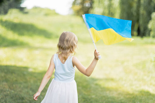 назад вид маленькой девочки с развевающимся сине-желтым флагом Украины, гуляющей по полю в солнечный летний день, концепция Дня Независимости Украины
