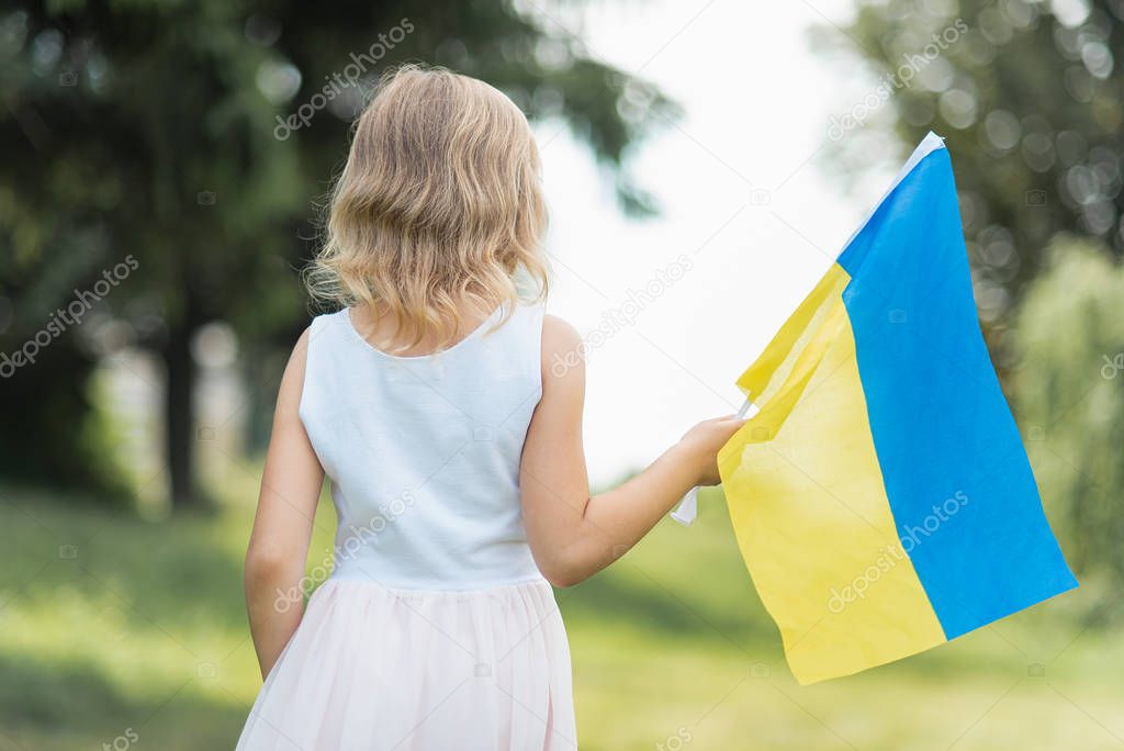 back view of little girl with fluttering blue and yellow flag of Ukraine walking in field at sunny summer day, Ukraine Independence Day concept
