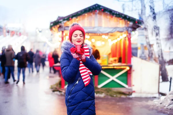 クリスマスマーケットでパンチを飲む若い女性 通りに立つ赤い帽子のかわいい女の子とホットワインや紅茶やカカオ 冬の雪の日に屋外のホットコーヒーを飲む クリスマスライト — ストック写真