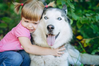 Parkta köpeğe sarılan sevimli küçük kız. Güzel güneşli hava, parlak güneş ışığı ve sevimli modeller. Pembe dili sarkmış, ağır ağır nefes alan bir köpek.