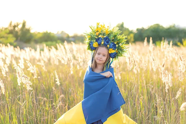 青いと黄色のウクライナの旗を保持し 夏の牧草地で歩くリボンと花の花輪を身に着けているかわいい女の子 ウクライナ独立記念日のコンセプト ウクライナの国旗の日 — ストック写真