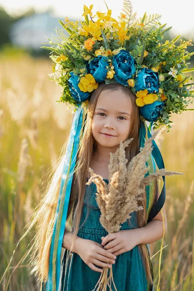 かわいい女の子は 青と黄色のリボンと花の花輪を身に着けていますウクライナの国旗の色小麦の小花の花束を保持し 夏の牧草地で歩く ウクライナ独立記念日の概念 ウクライナの国旗の日 — ストック写真