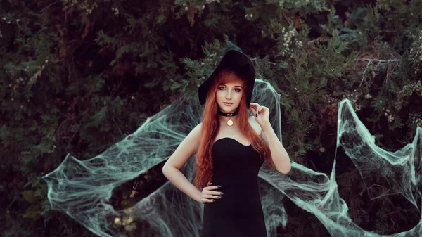 ハロウィンの肖像画を閉じる不気味な暗い魔法の森の背景に魔女の帽子の美しい若い魔女の女の子 大きな目の魔法の光景を持つ赤毛の女の子 — ストック写真
