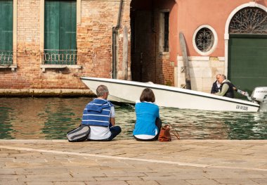 Venedik, İtalya - 13 Ocak 2019: Bir çift turist önlerinde bir motorlu tekne seyrederken bir kanalın kenarında oturuyorlar. Venedik, UNESCO dünya mirası sahası, Veneto, İtalya, Avrupa