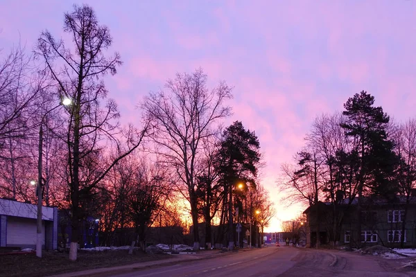 小さな町の早朝の紫の空。明かりが燃えている空っぽの道ドラマチックな夕日の背景. — ストック写真