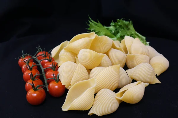 Pasta sotto forma di conchiglie in una ciotola di legno rotonda con pomodorini su sfondo nero. Contesto culinario . — Foto Stock