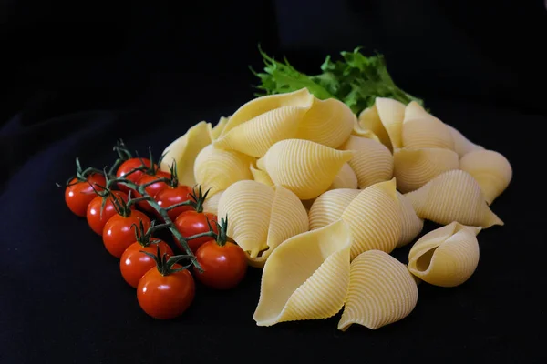 Pasta in de vorm van schelpen in een ronde houten schaal met cherry tomaten op een zwarte achtergrond. Culinaire achtergrond. — Stockfoto
