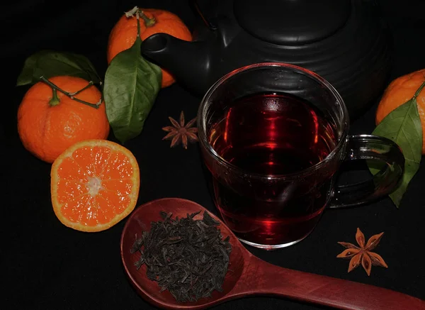 Thé d'automne dans une tasse transparente. Couper les mandarines sur un fond noir. Thé dans une cuillère en bois — Photo