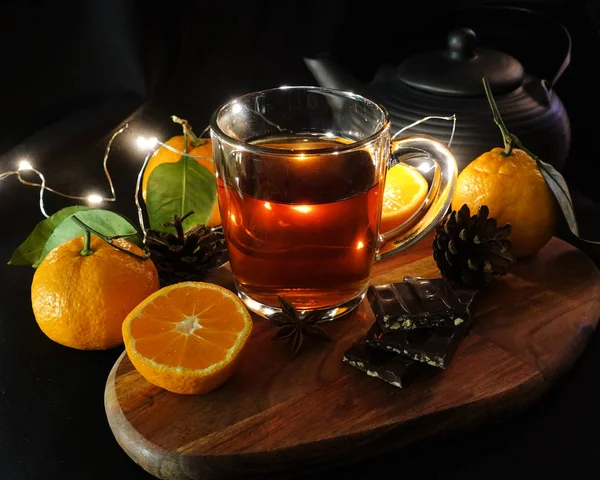 Autumn tea in a transparent cup. Cut tangerines on a black background. Broken chocolate. Garland. Black background