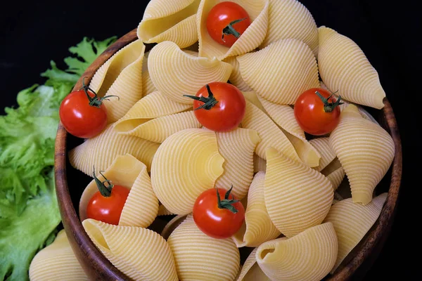 Makaron w postaci muszli w okrągłej drewnianej misce z pomidorami wiśniowym na czarnym tle. Tło kulinarne. — Zdjęcie stockowe