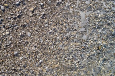 Deniz plajında küçük çakıl taşları ve ıslak kum; su hattı. Sakin şeffaf su.