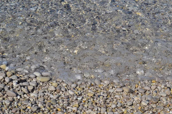 Deniz plajında küçük çakıl taşları ve ıslak kum; su hattı. Sakin şeffaf su. — Stok fotoğraf