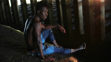 Afrika delikanlı ile çıplak göğüslü Jeans kaya beton destekler arka plan üzerinde köprü altında oturur.