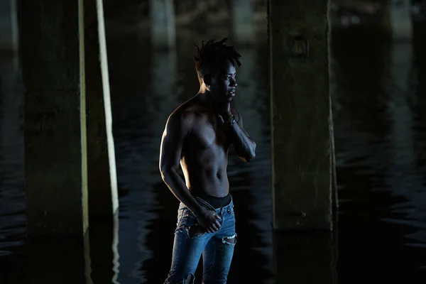 Young African man with bare-chested in jeans stands in water under bridge on background of concrete supports.