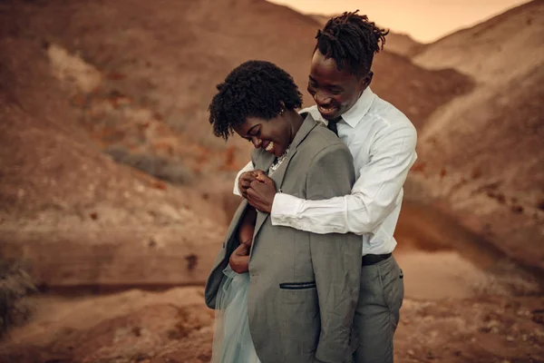 Black groom hugs his bride dressed in his jacket in canyon against beautiful landscape at sunset. They laugh cheerful. Closeup.