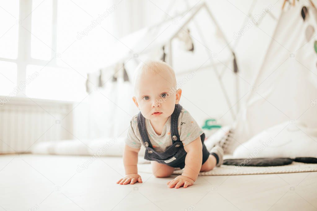 Toddler in rompers and t-shirt crawls on floor in room against window.