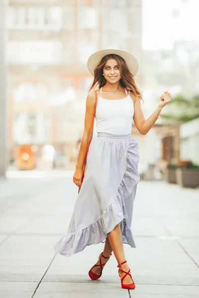 Young woman in hat and long skirt walks along city street.