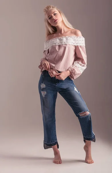 Blonde barefoot model in pink blouse with lace and jeans is posing in studio on pink background.