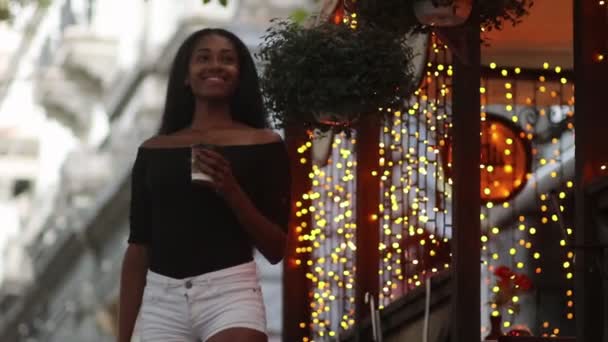 年轻女子走在街上 喝咖啡从杯子和微笑的背景下的城市灯光晚上 散景效果 — 图库视频影像