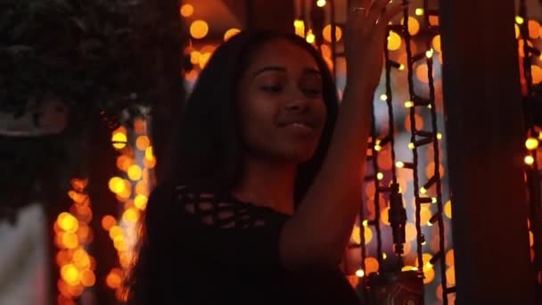 若い幸せな女は 店の窓の隣に立っているし 夜の街の明かりの背景に手をガーランドに触れます ピンぼけ効果 — ストック動画