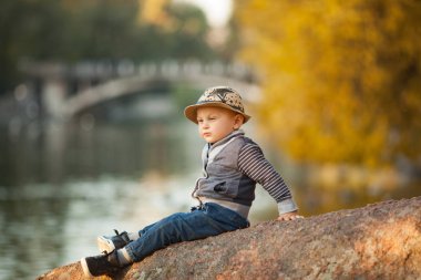 Küçük bir çocuk bir kayaya göl yanındaki sarı sonbahar ağaçlar arka planı parkta bir yürüyüş sırasında oturuyor.