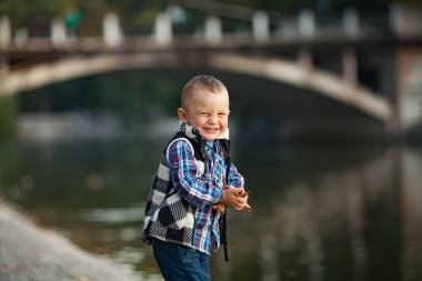 Küçük bir çocuk oyun ve parkta bir yürüyüş sırasında bir göl ve bir köprü arka planı elinde sonbahar yaprakları tutarak. Neşeyle gülüyor.