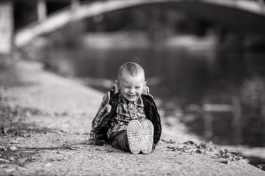 Küçük bir çocuk, parkta bir yürüyüş sırasında göl ve bir köprü arka planı sonbahar yaprakları yanında oturuyor. Neşeyle gülüyor. Siyah beyaz resim.