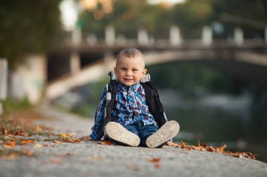 Küçük bir çocuk parkta bir yürüyüş sırasında sarı sonbahar yaprakları göl ve bir köprü arka planı yanında oturuyor.