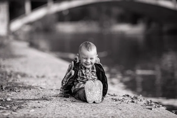 Küçük bir çocuk, parkta bir yürüyüş sırasında göl ve bir köprü arka planı sonbahar yaprakları yanında oturuyor. Neşeyle gülüyor. Siyah beyaz resim.
