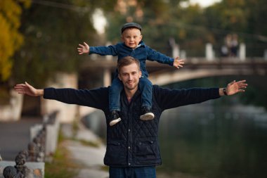 Genç bir baba küçük oğlu ile göl ve köprü arka planı parkta bir yürüyüş sırasında onun omuzlarında duruyor.