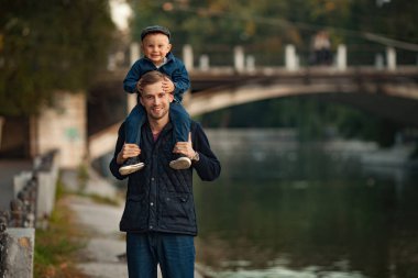 Genç bir baba küçük oğlu ile göl ve köprü arka planı parkta bir yürüyüş sırasında onun omuzlarında duruyor.