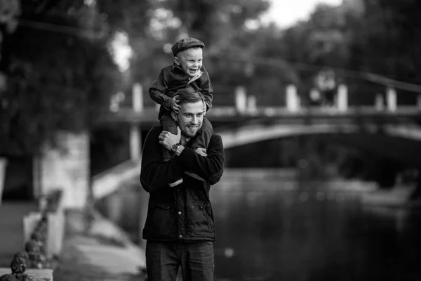 若い父親は湖と橋の背景に公園を散歩中に彼の肩に彼の幼い息子と立っています 黒と白のイメージ — ストック写真