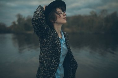 Akşamları sırasında alacakaranlık gökyüzü arka plan üzerinde göl yanındaki şapkalı genç kadın standları.