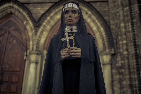 Beautiful young nun in black cassock, cloak and transparent cape is standing and praying with cross in her hands on temple background. Closeup.
