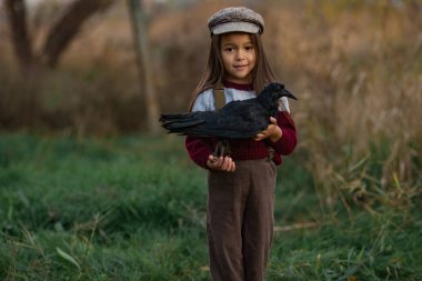 Şapkalı kız çocuk elleri yeşil çim ve ağaçların arka plan üzerinde siyah Kuzgun ile duruyor.