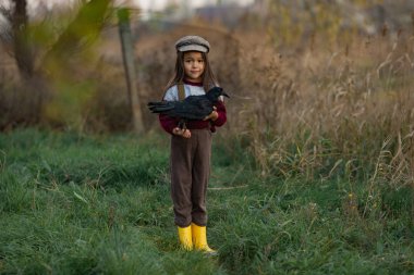 Şapkalı kız çocuk elleri yeşil çim ve ağaçların arka plan üzerinde siyah Kuzgun ile duruyor.
