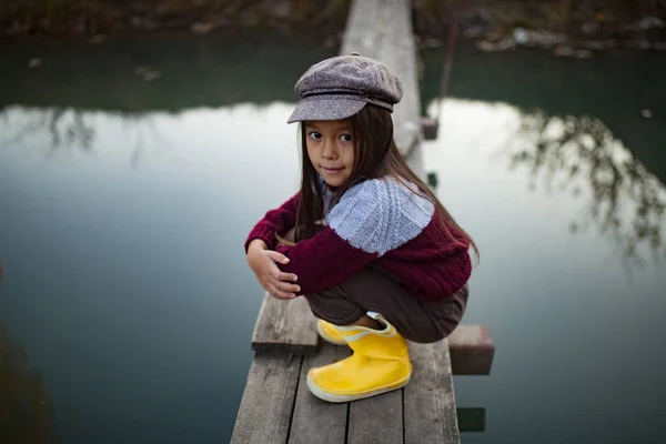 Child girl in cap and yellow rubber boots sits on wooden bridge on background of river.