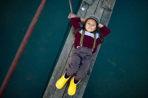 Child girl in cap and yellow rubber boots lies on wooden bridge and smiles on background of river. Top view.