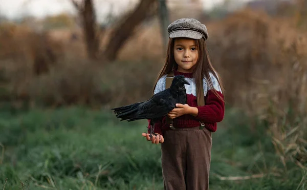 戴帽子的女孩站在她的手中的绿色草地和树木的背景黑色乌鸦 — 图库照片
