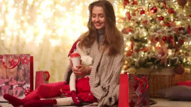 Ekose ve Noel Baba ile genç kadın oturur yanındaki Noel ağacı ve hediye paketleri, gülümsüyor ve karton bardak köpüklü Işık Ampüller arka plan üzerinde kahve içer. 