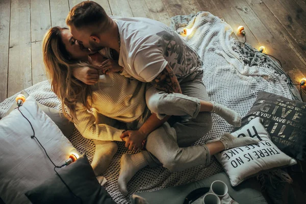 一对恋爱中的年轻夫妇在木地板 枕头和发光的灯泡的背景下拥抱和亲吻 — 图库照片