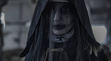 Portrait of standing in a darkness woman dressed in a black hooded cloak in an image of a nun possessed by demons. Cosplay. clipart