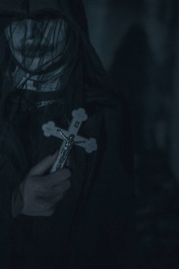 Kadın bir karanlıkta bir haç ile bir rahibe iblisler tarafından ele geçirilmiş bir görüntüdeki siyah bir kukuletalı pelerin elinde duruyor. Cosplay.