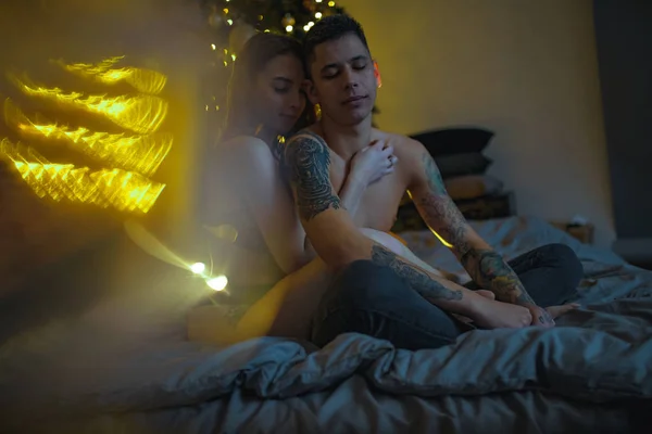 クリスマス ツリーや装飾品の背景にベッド上で抱擁で恋に半分裸のカップルが座っています カーテンの隙間から表示します — ストック写真