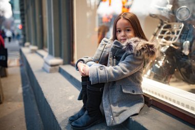 Bir ceket kürk başlık ile kız çocuk akşamları caddenin üzerinde shopwindow yanında oturuyor. Portre.