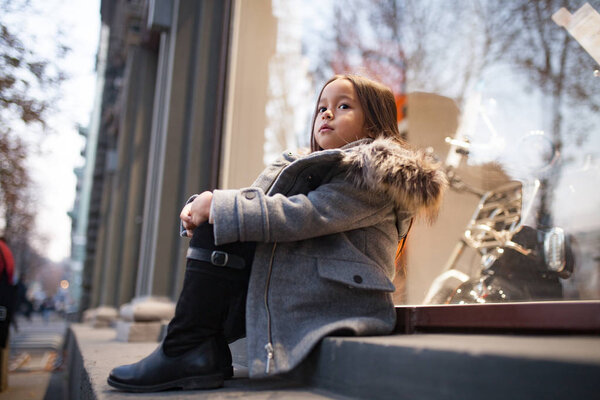 Child girl in a coat with a fur hood is sitting next to shopwindow on city street in evening. Closeup.