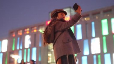 Bir ceket, şapka ve atkı, kız çocuk döne döne ve şehir ışıkları akşam arka plan karşı Street onun elinde smartphone ile kendini fotoğrafını çekmek.
