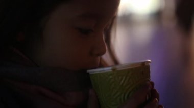 Bir ceket, şapka ve atkı, kız çocuk ayakta ve Street insanlar tarafından geçen ve şehir ışıkları akşam arka plan karşı onun elinde tek kullanımlık Kupası Buhar ile sıcak kahve içme.