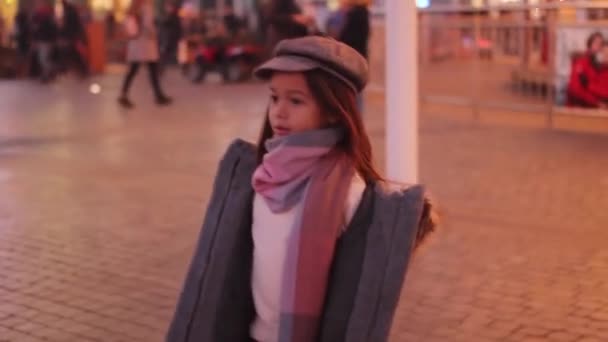 穿着外套 帽子和围巾的女孩在经过人们和傍晚城市灯光的背景下走在大街上 — 图库视频影像